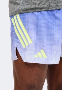 Gros plan d'une personne portant un short de sport violet clair avec des bandes jaune pâle et un petit logo triangulaire sur la cuisse gauche.