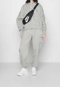 Osoba oblečená v svetle šedé mikině Nike a cargo kalhotách s bílými teniskami, nese černou crossbody tašku Nike.