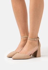 Beige kärjelliset korkokengät, joissa on lohkokorko ja nilkkaremmi. Valmistettu sileästä materiaalista, minimalistisella muotoilulla ja tukevalla suljinmekanismilla.