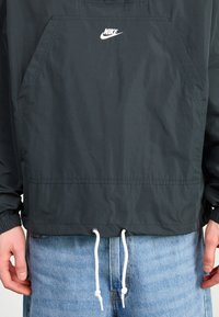 Coupe-vent noire Nike avec logo blanc, poche kangourou à l'avant et cordons blancs, porté sur un jean en denim bleu clair.