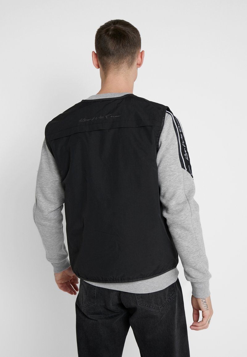 Svart nylonväst över en grå långärmad topp, med ett subtilt sömmönster och svart-vita randiga detaljer på ärmarna.