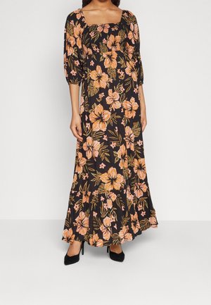 Vrouw draagt een zwarte maxi-jurk met oranje bloemenprint en opgepofte driekwartsmouwen, gecombineerd met zwarte hoge hakken.