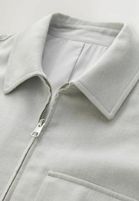 Veste gris clair avec un col pointu, fermeture éclair argentée partiellement ouverte et une poche avant visible sur un tissu texturé.