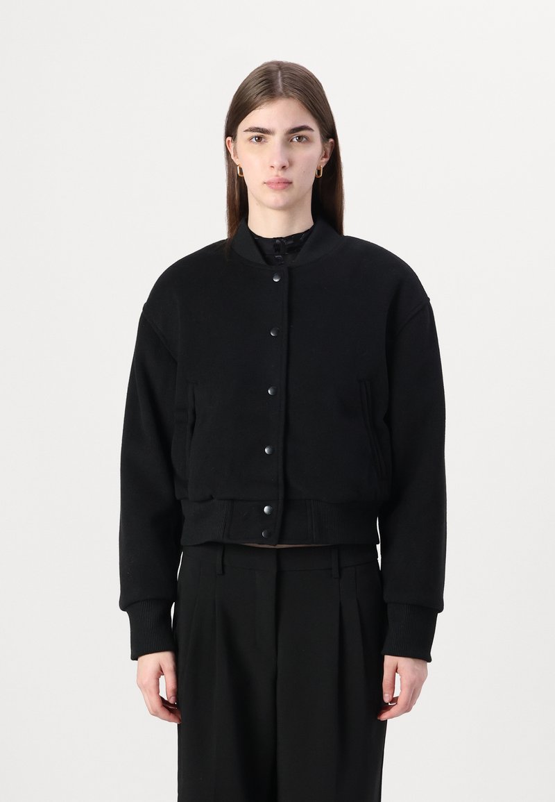Giacca bomber cropped nera con bottoni a pressione, polsini a costine e tasche laterali. Realizzata in un tessuto morbido con una texture liscia.