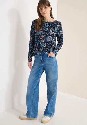 Vrouw die een blauw, bloemig topje met lange mouwen, wijde blauwe jeans en witte sneakers draagt, staand tegen een tweekleurige beige-witte muur.