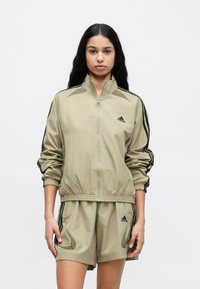 Svetlokhaki športová bunda so zipsovým zapínaním, elastickými manžetami a čiernymi bočnými pruhmi. Kombinovaná s rovnako farebnými šortkami s logom a textúrovanými detailmi.