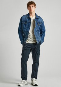 Jeans jakna, prugasta majica in temni cargo hlače. Ima žepke, klasičen kroj in sproščen fit. Tenisice dopolnjujejo videz.
