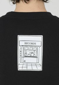 T-shirt noir avec une illustration imprimée blanche d'un magasin de disques, avec une guitare et des percussions visibles dans la vitrine.