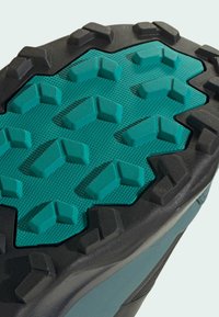 Trail-Schuhsohle mit einer strukturierten, schwarzen Gummirand und einem türkisfarbenen, gemusterten Griffbereich mit eckigen Stollen für besseren Halt.