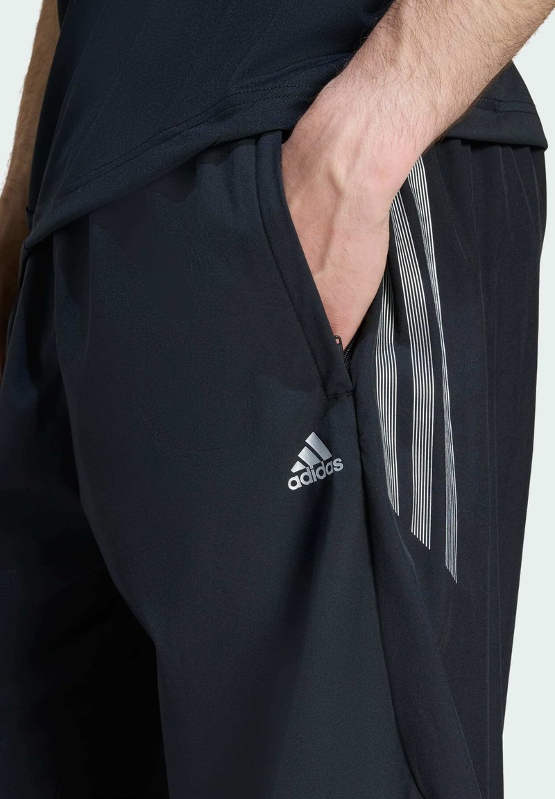 Svarta träningsshorts med en ficka med dragkedja, tillverkade av ett texturerat tyg och tre vertikala vita ränder på sidan. Adidas-logotyp på fållen.