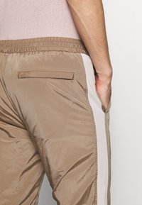 Pantalones deportivos color marrón claro hechos de tela suave y brillante con una cintura elástica y una franja lateral blanca. Incluyen un pequeño bolsillo trasero.