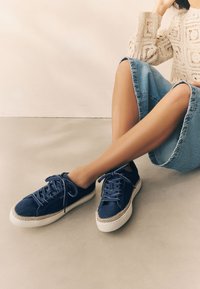 Marineblå lærredssneakers med hvide gummisåler, med kontrasterende snøredetaljer og en tekstureret tåhætte, stylet med et denimbuks.