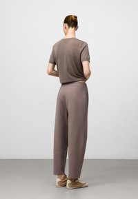 Vrouw met haar in een knot, gekleed in een mauve kortemouwen top en bijpassende wijde broek, staand met haar rug naar de camera tegen een effen achtergrond.