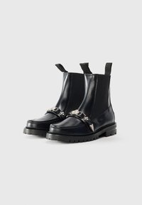 Bottines en cuir noir avec des panneaux latéraux élastiques, semelle en caoutchouc épaisse, accents argentés et boucles décoratives à l'avant.