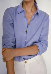 Ljusblå skjorta med lång ärm som är uppvikt vid ärmsluten. Mjukt tyg med en subtil textur. Matchad med vita byxor med en blank knapp.