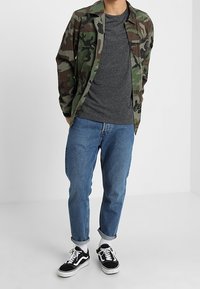 Kamouflagemönstrad jacka i grön och brun, grå t-shirt, blå jeans och svarta sneakers med vita detaljer. Avslappnad passform och upprullade ärmar.