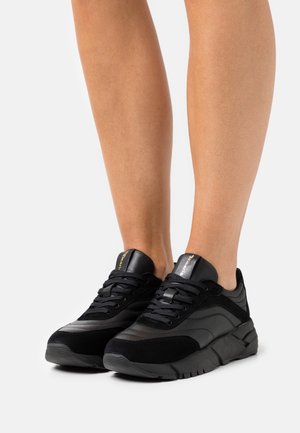 Zapatilla negra con una mezcla de texturas lisas y de ante, con suela sólida, cuello acolchado y cordones negros. Marca en dorado en la lengüeta.