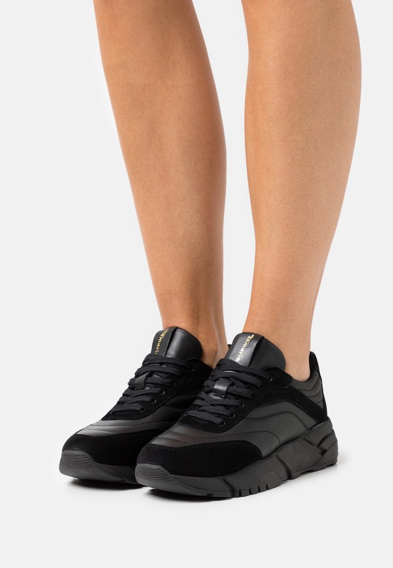 Schwarze Sneaker mit einer Mischung aus glatten und Wildledertexturen, ausgestattet mit einer stabilen Sohle, gepolstertem Kragen und schwarzen Schnürsenkeln. Goldene Markierung auf der Zunge.