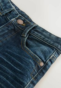 Vaqueros de mezclilla en azul oscuro con costuras tonales, botón metálico y diseño de cinco bolsillos. Textura desvanecida a lo largo de la pierna y la cintura.