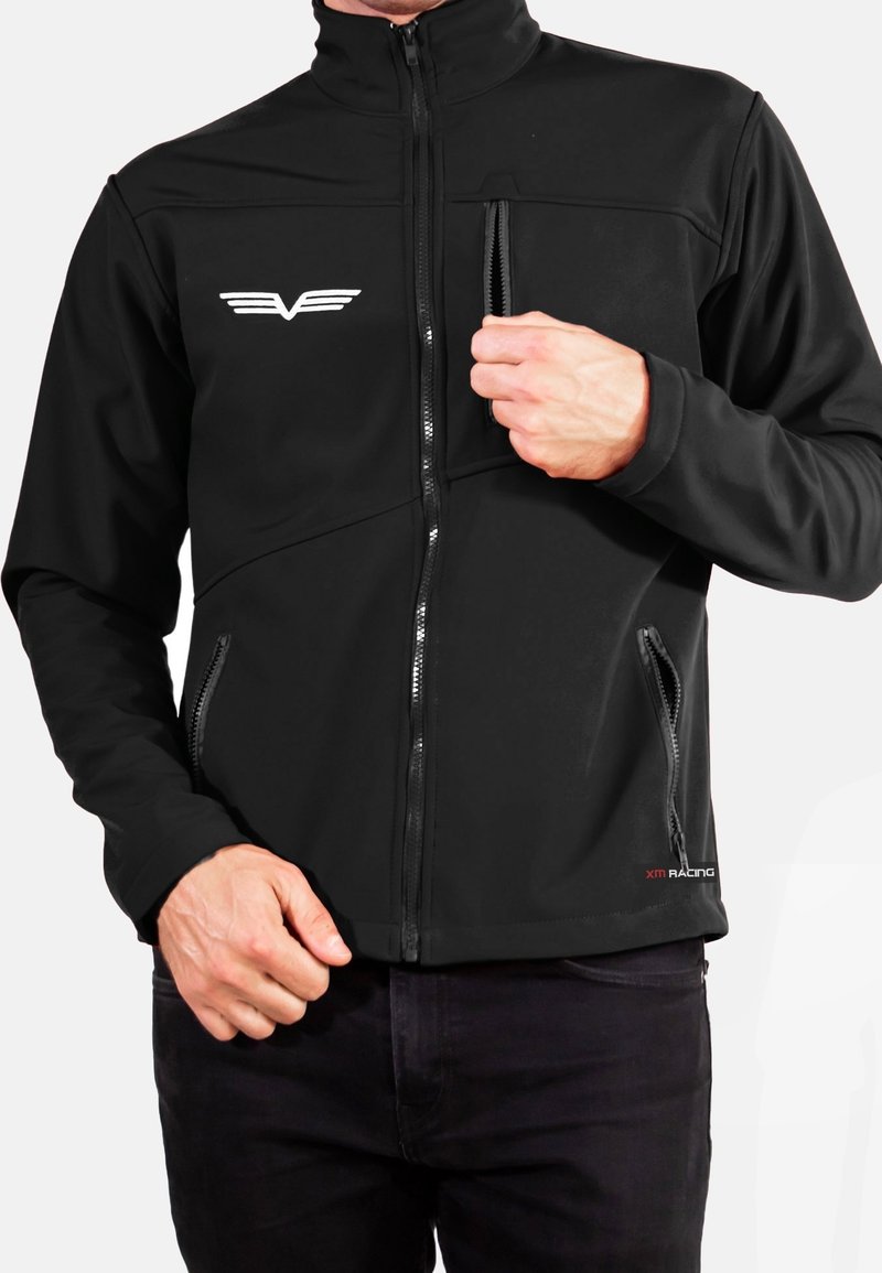 Veste softshell noire avec un col montant, des poches zippées et un emblème sur le devant. Présente une texture lisse et une coupe ajustée.