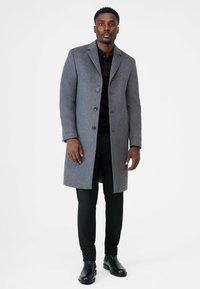 Grijze wollen jas met een geknipt kraag, drie knopen en zijzakken. Gecombineerd met een zwart shirt en zwarte broek, aangevuld met glanzende laarzen.