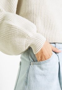 Hand i en ljusblå denimjeansficka, iklädd en krämfärgad grovstickad tröja med ribbade manschetter.