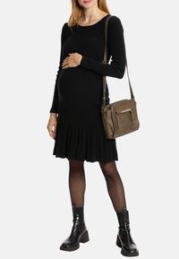 Schwarzes langärmeliges Kleid mit plissierter Rockpartie, gefertigt aus weichem Stoff. Das Model trägt eine bronzefarbene Handtasche und schwarze Stiefeletten.