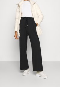 Persona che indossa pantaloni neri a gamba larga con coulisse, top bianco a costine, giacca puffa color crema e sneaker chunky bianche su uno sfondo grigio.
