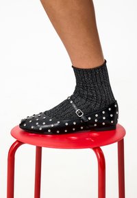 Scarpa piatta nera in pelle con dettagli a borchie argentate, cinturino con fibbia, indossata con un calzino grigio a coste, posizionata su uno sgabello rosso.