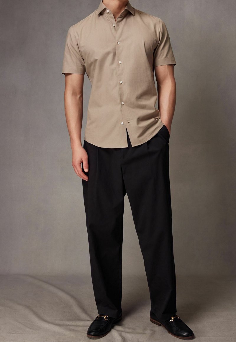Homme portant une chemise beige à manches courtes boutonnée, un pantalon noir ample et des mocassins noirs, debout avec une main dans la poche.
