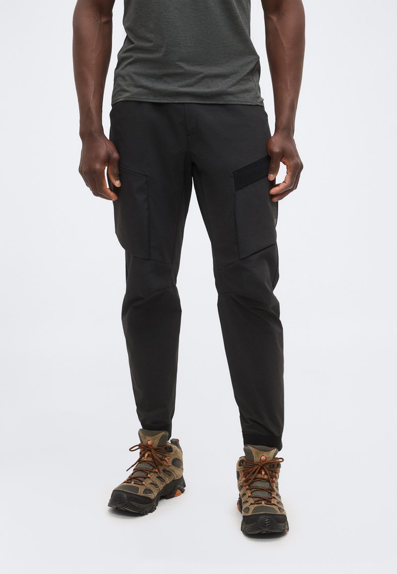 Άνδρας που φοράει μαύρο παντελόνι cargo με πολλές τσέπες και καφέ ορειβατικά παπούτσια, στέκεται μπροστά σε ένα απλό φωτεινό φόντο.