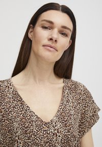 Top con stampa animale caratterizzato da scollatura a V, maniche corte, sfondo beige con macchie marroni e nere, tessuto testurizzato, design casual.