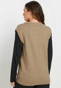 Gilet lavorato a maglia beige con colletto rotondo e design senza maniche, indossato sopra una maglietta nera a maniche lunghe. Sono visibili fessure laterali e una texture morbida.