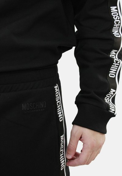 Tuta nera Moschino con stampa del logo bianco lungo le maniche e i pantaloni, con polsini a costine e logo impresso sulla vita.