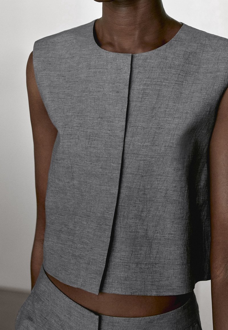 Haut court gris texturé sans manches avec une couture centrale à l’avant, porté par une personne à la peau foncée.