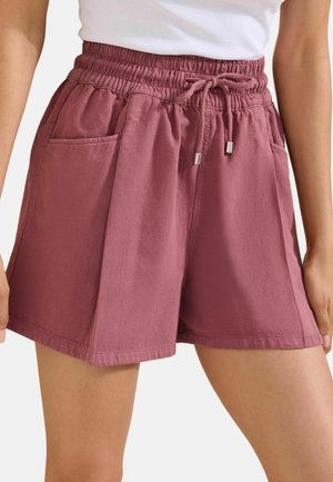 Frau trägt hoch taillierte Shorts in Mauve mit elastischem Bund, Kordelzug und vorderen Taschen, kombiniert mit einem weißen ärmellosen Top.