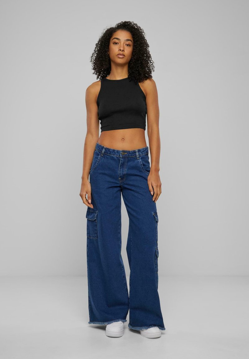 Svart kort crop-top kombinerad med lösa, blå denim-cargo-byxor med sidofickor och fransade fållar; modellen bär vita sneakers.