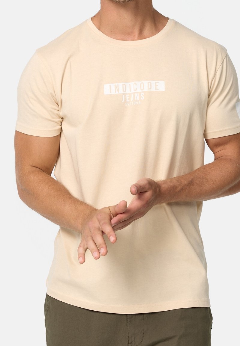 Homme portant un t-shirt beige à manches courtes avec le texte « INDICODE JEANS », ajustant ses mains devant un fond gris clair.