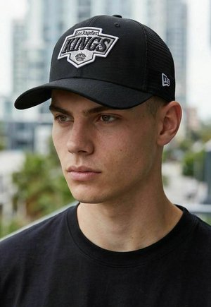 Junger Mann mit einer schwarzen Los Angeles Kings Cap und schwarzem Hemd, vor einem verschwommenen städtischen Stadtbild im Hintergrund.