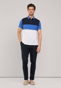 Polo shirt in blauw, marineblauw en witte kleurvlakken; korte mouwen; kraag; gedragen met donkere broeken en beige schoenen; casual pasvorm.