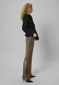 Schwarzer Blazer über einem taillierten, schimmernden goldenen Rock mit seitlichen Schlitzen. Das Outfit wird mit schwarzen spitzen Pumps und einer eleganten Silhouette kombiniert.