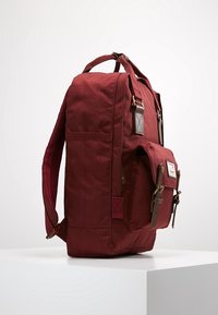 Bordowy plecak z płótna z brązowymi skórzanymi akcentami, regulowanymi paskami i zewnętrzną kieszenią. Wyróżnia się teksturowanym materiałem i solidnymi elementami metalowymi.