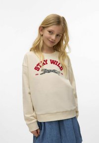 Blond tjej i en krämfärgad sweatshirt med texten "STAY WILD" och en grafisk bild av en springande hund, i kombination med en blå kjol, leende lätt.
