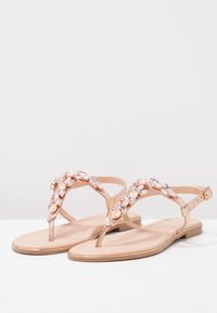 Beige litte sandaalit, joissa on T-nauhat, jotka on koristeltu vaaleanpunaisilla ja kirkkailla jalokivikoristeilla sekä säädettävillä nilkkahihnoilla.