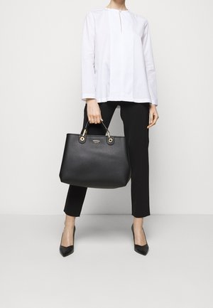 Bolso tote de cuero negro con herrajes dorados, que cuenta con dos asas superiores y una superficie texturizada, combinado con una blusa blanca y pantalones negros.