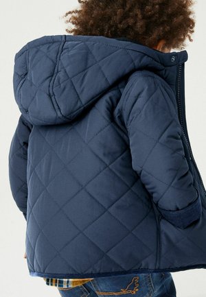 Veste matelassée marine avec capuche, présentant un motif texturé en losanges, des poignets côtelés et une fermeture éclair. Portée sur une chemise à carreaux.