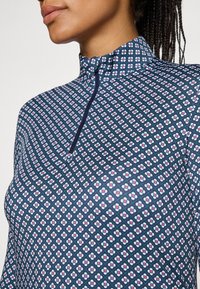 Langärmliges Oberteil mit hohem Kragen, aus einem dehnbaren Stoff mit einem geometrischen blau-weißen Blumenmuster und einem Reißverschluss vorne.