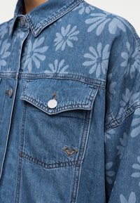 Μπλε τζιν μπουφάν με φωτεινά γαλάζια λουλουδάτα σχέδια, μεταλλικά κουμπιά, τσέπη με καπάκι και μικρό λογότυπο σε σχήμα καρδιάς στο στήθος.