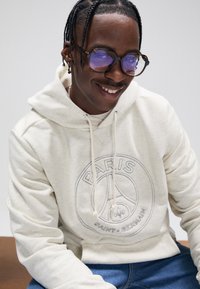 Sweatshirt com capuz cor creme, com um logótipo gráfico circular que diz "PARIS SAINT-GERMAIN". Apresenta cordões para ajuste e um tecido macio e texturizado.