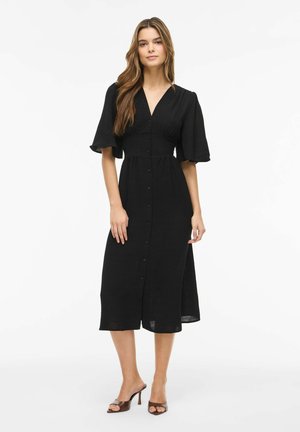 Mujer con vestido midi negro con botones delanteros, escote en V y mangas cortas y anchas, combinada con sandalias marrones de tacón abierto.